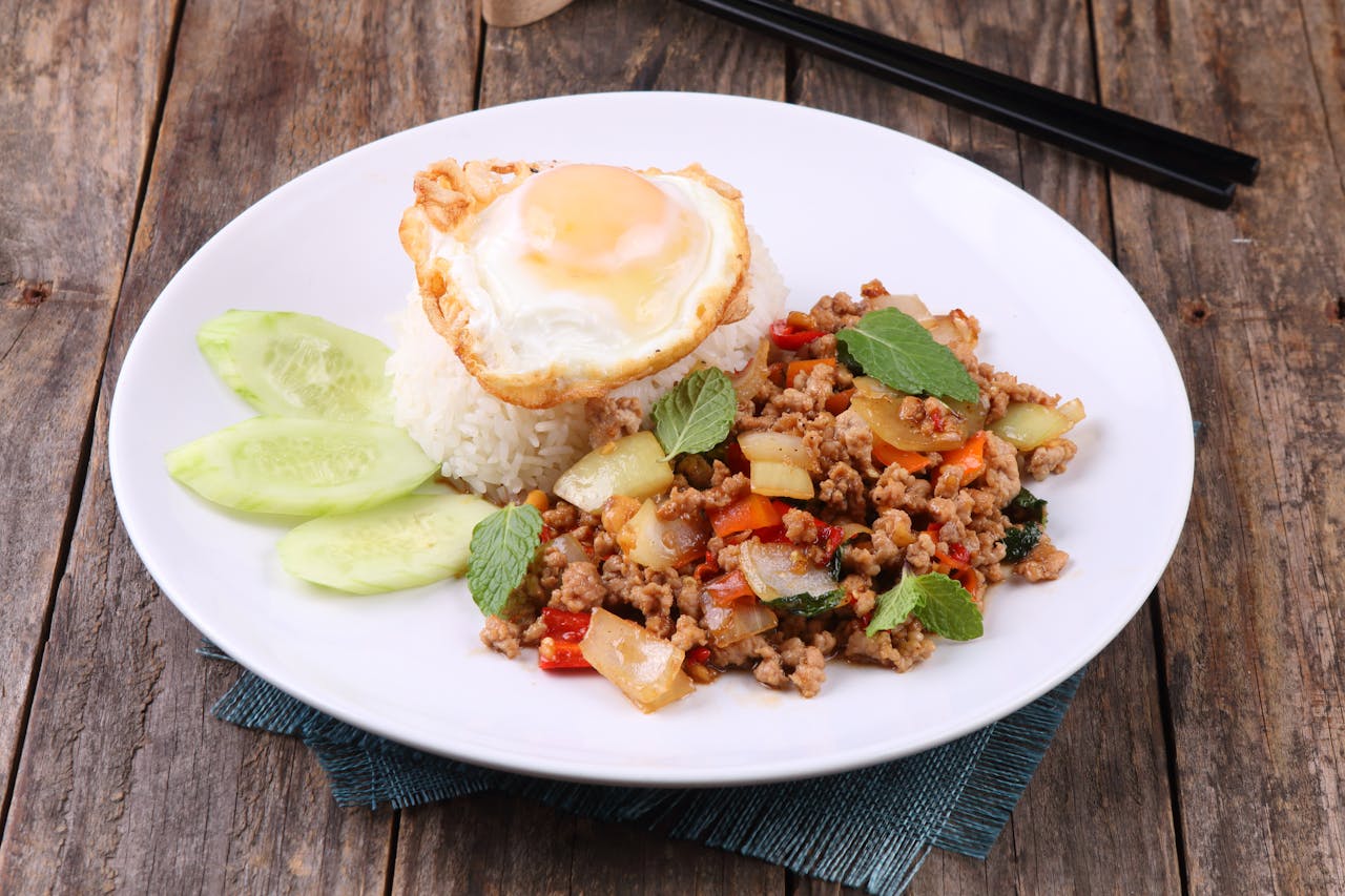 Thai stir-fry dish with rice and egg on rustic table setting, vibrant and appetizing.